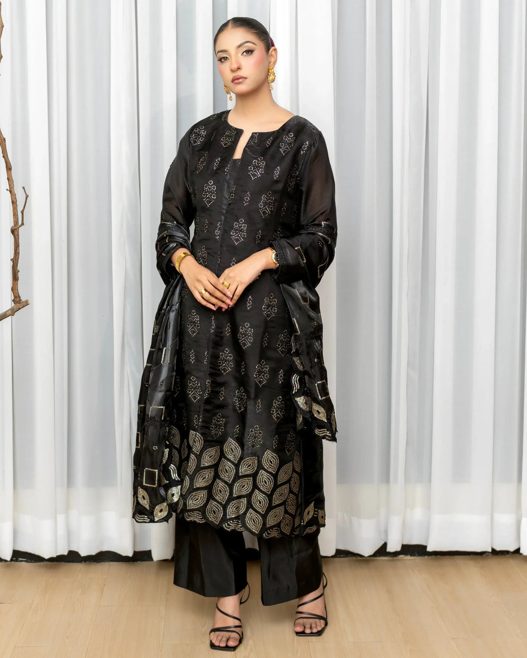 Woman wearing a black patterned outfit standing against a white curtain.