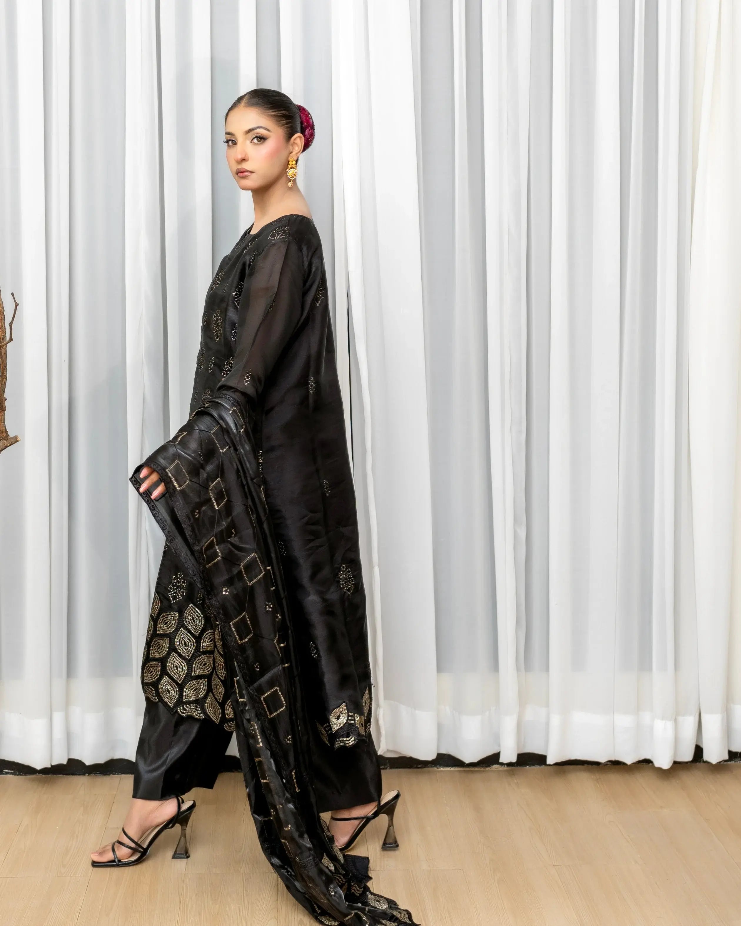 Woman in a black outfit with intricate patterns standing against a white curtain.