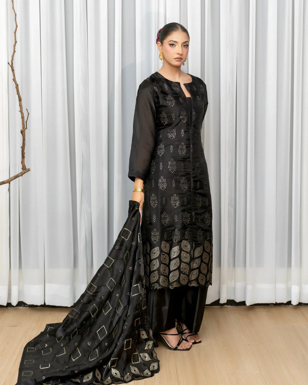 Woman in a black traditional outfit with a long scarf against a white curtain background