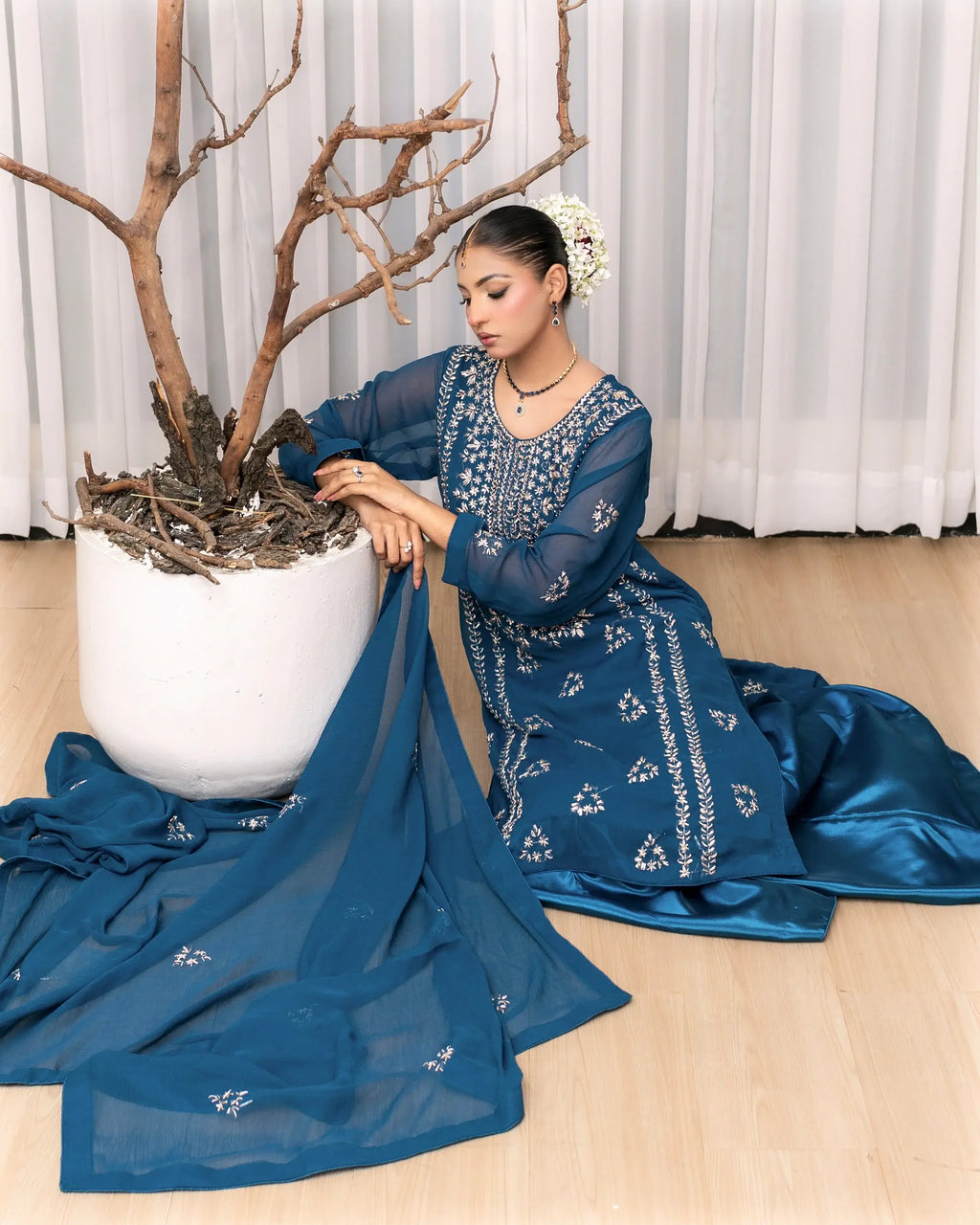 Woman in a blue traditional outfit sitting next to a white pot with branches indoors.