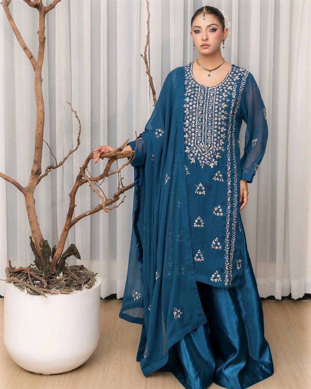 Woman wearing a blue embroidered traditional outfit standing next to a plant indoors.