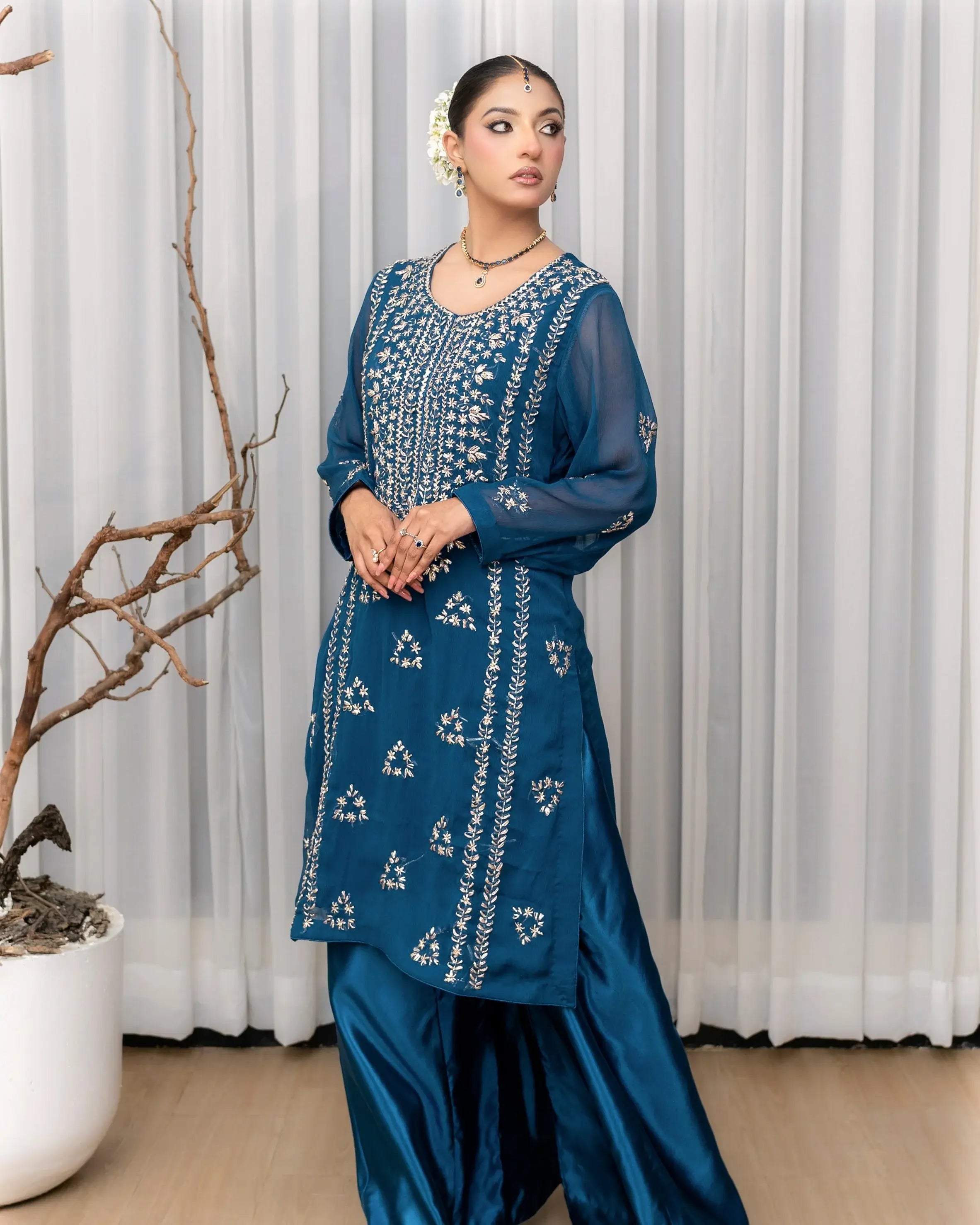 Woman wearing a blue embroidered traditional outfit standing against a white curtain.