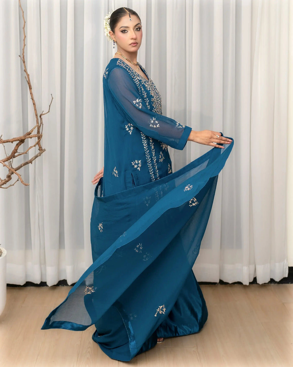 Woman wearing a blue saree with white embroidery against a white curtain background
