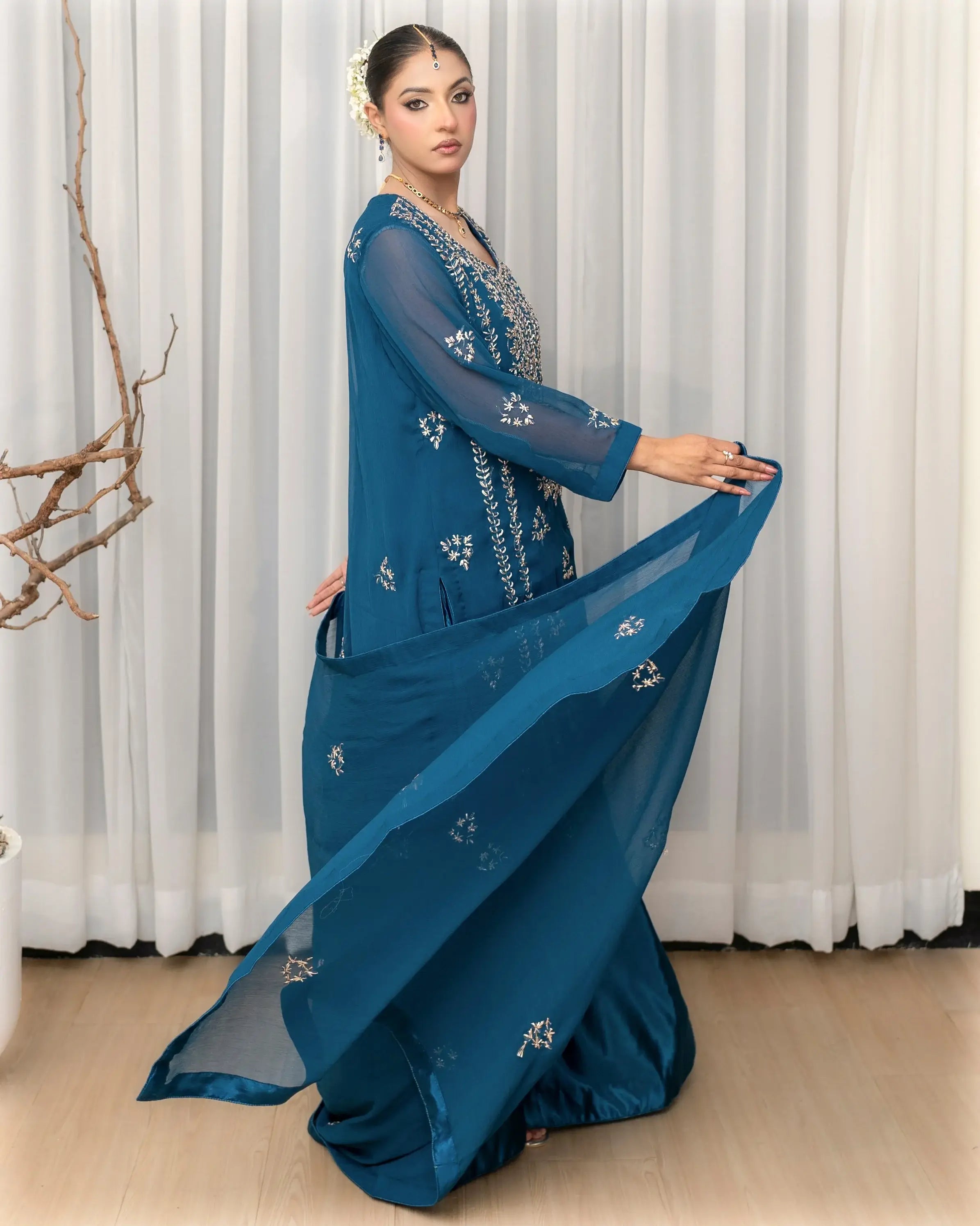 Woman wearing a blue saree with white embroidery against a white curtain background