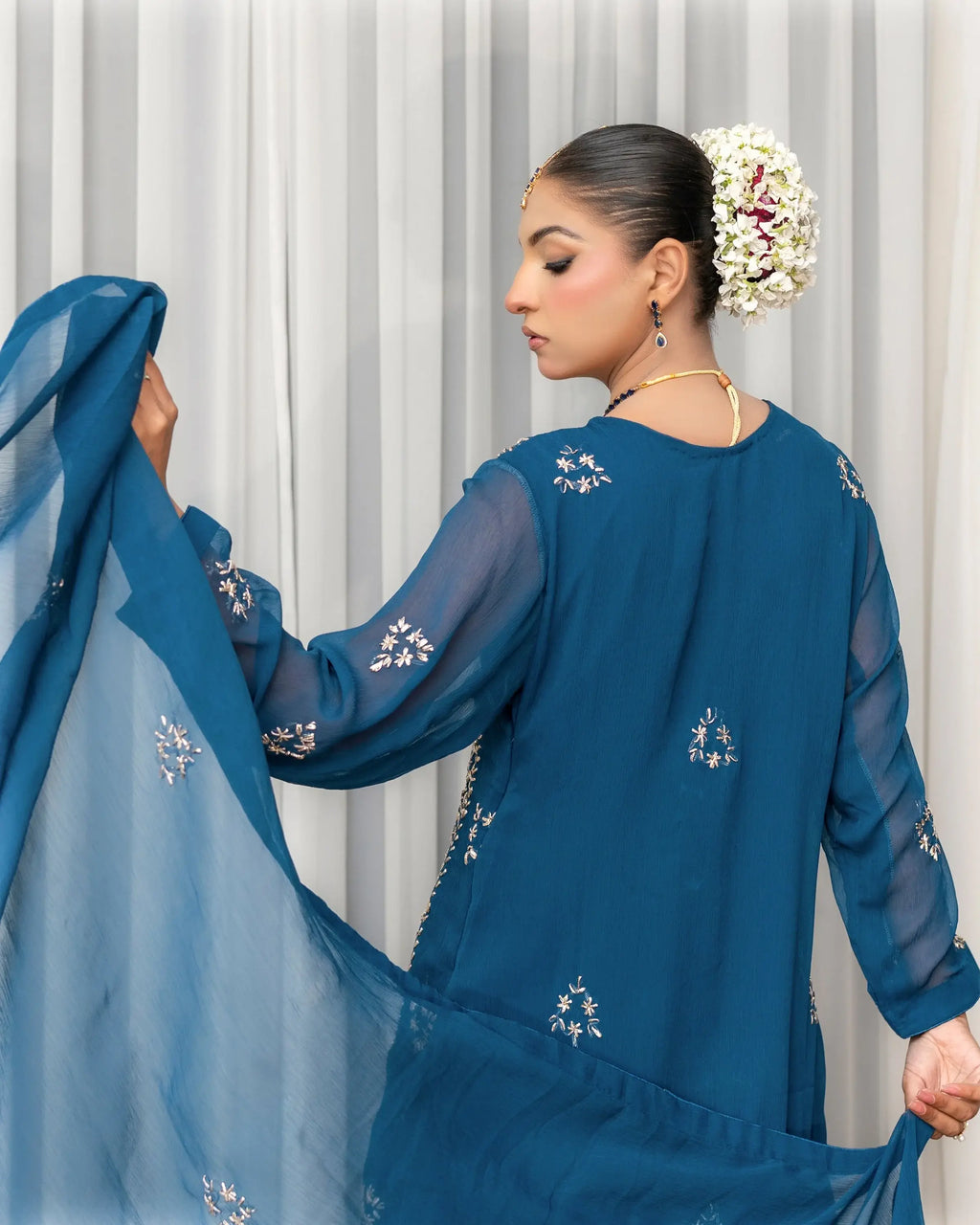 Woman wearing a blue embroidered outfit with a sheer overlay against a white curtain background