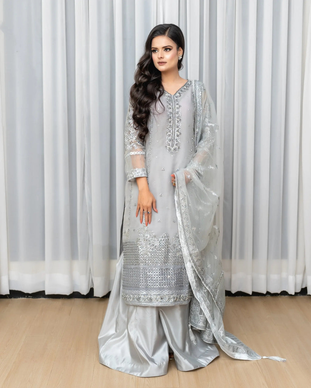 Woman wearing a light gray embroidered traditional outfit with a white curtain background