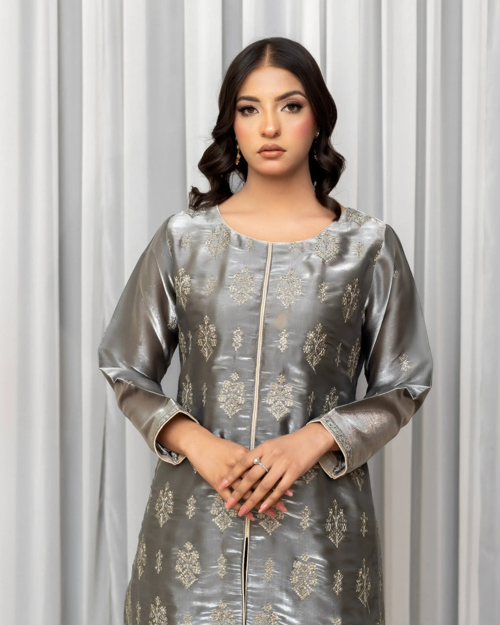 Woman wearing a silver traditional outfit with intricate patterns against a white curtain background