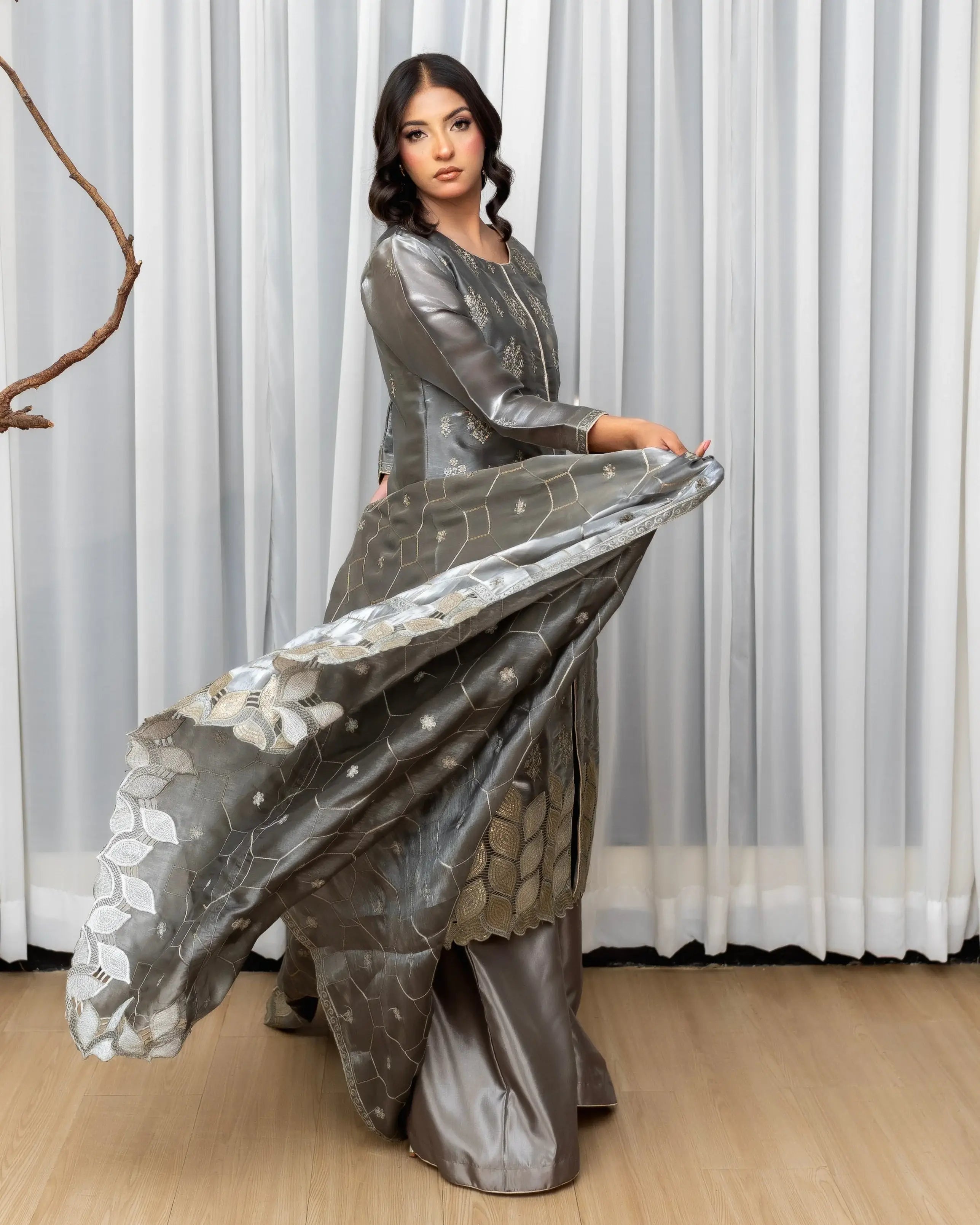Woman in a silver dress with a white floral pattern, standing against a white curtain.