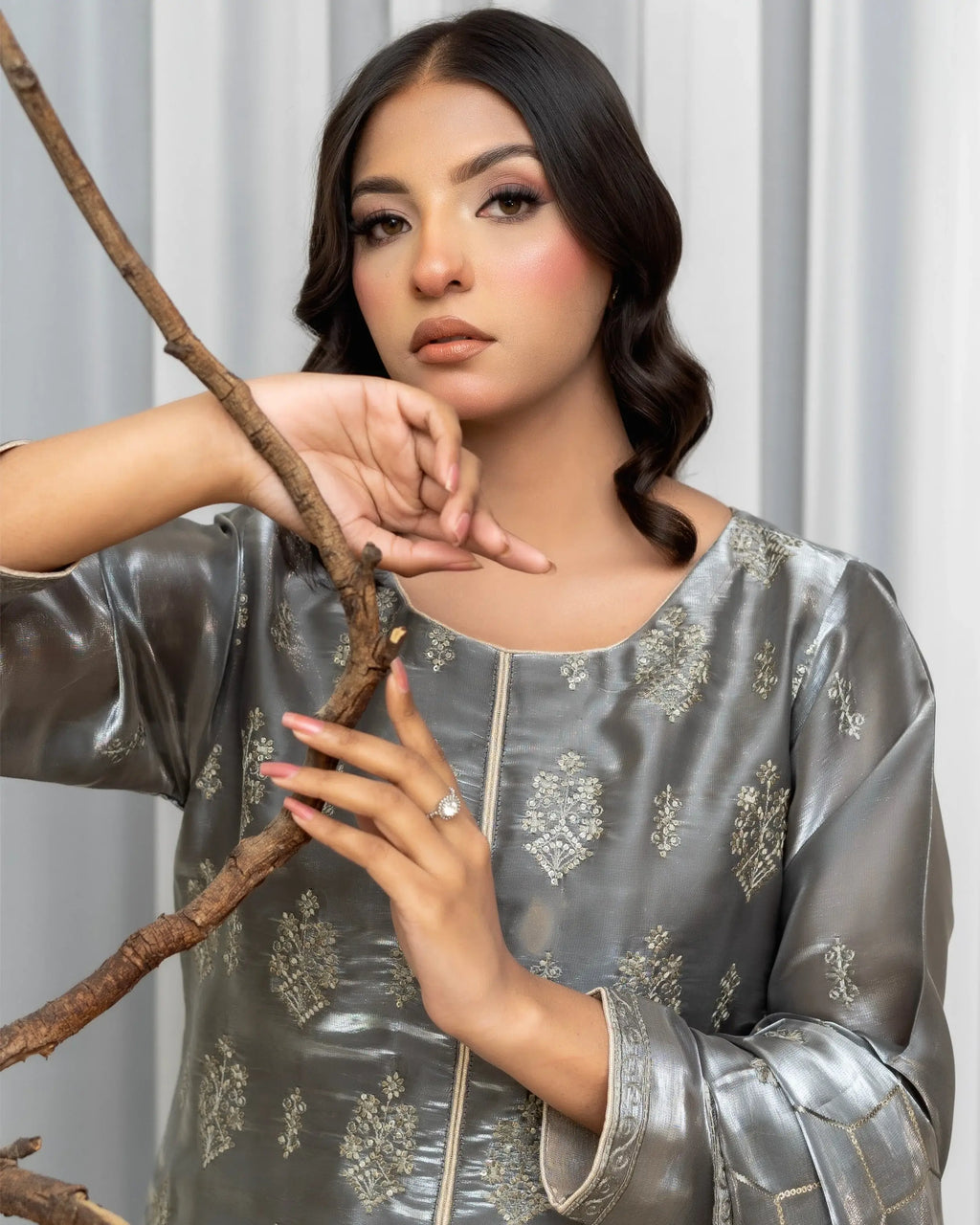 Woman in a silver traditional outfit holding a branch against a white curtain background