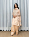 Woman wearing a beige traditional outfit with intricate patterns against a white curtain background