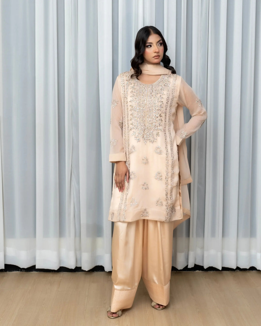 Woman wearing a beige traditional outfit with intricate patterns against a white curtain background