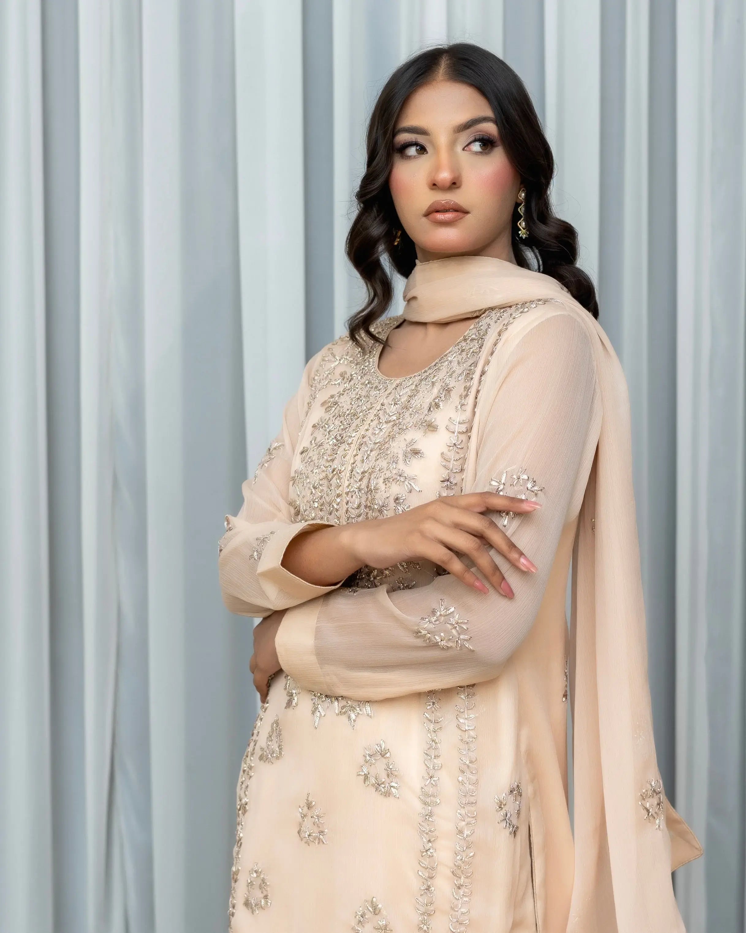 Woman wearing a beige embroidered traditional outfit against a light gray curtain background