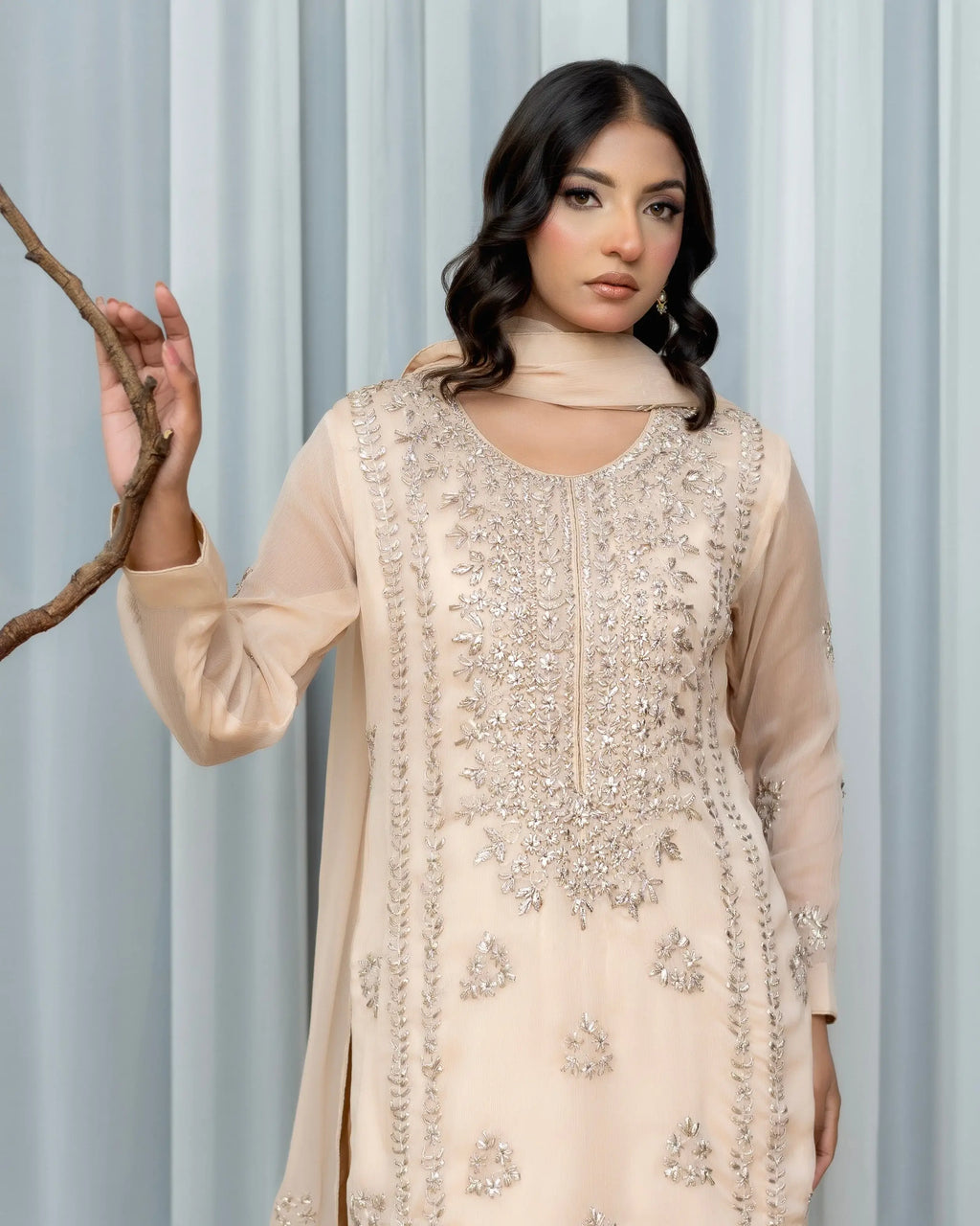 Woman wearing a beige embroidered traditional outfit against a light gray curtain background