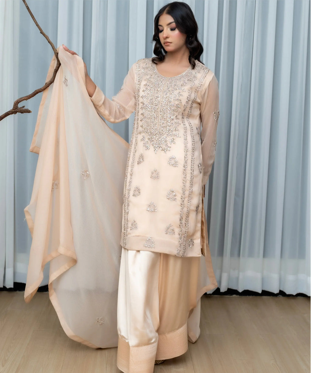 Woman in a beige embroidered traditional outfit holding a dupatta against a light curtain background