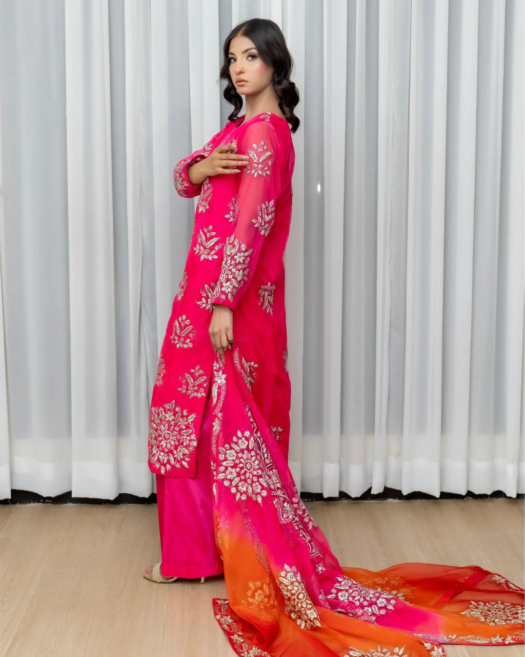 Woman wearing a pink and orange traditional outfit with floral patterns in front of white curtains.