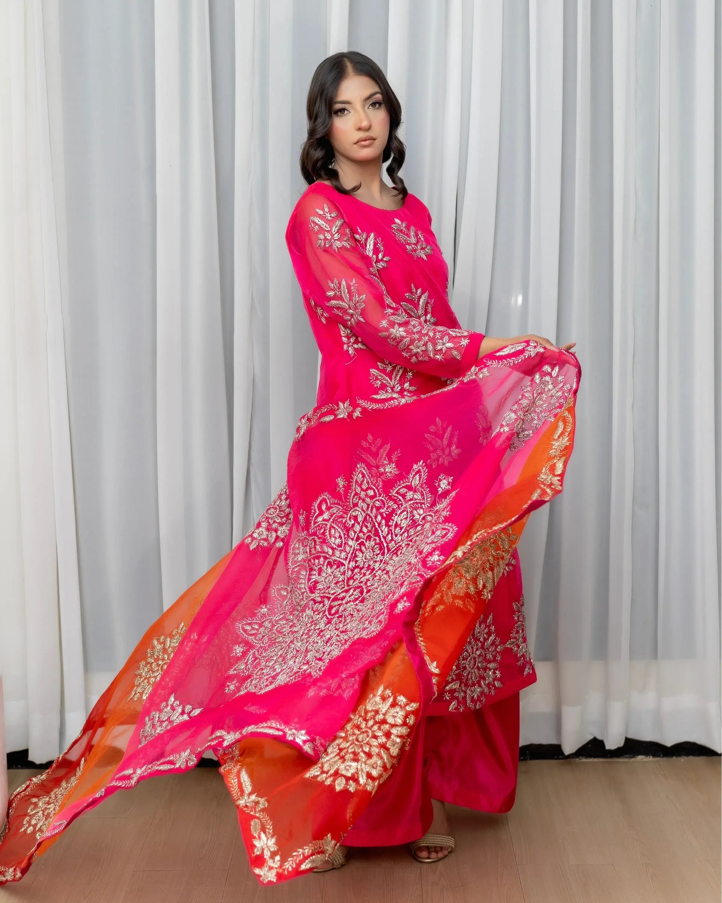 Woman wearing a pink and orange traditional outfit with white patterns against a white curtain background