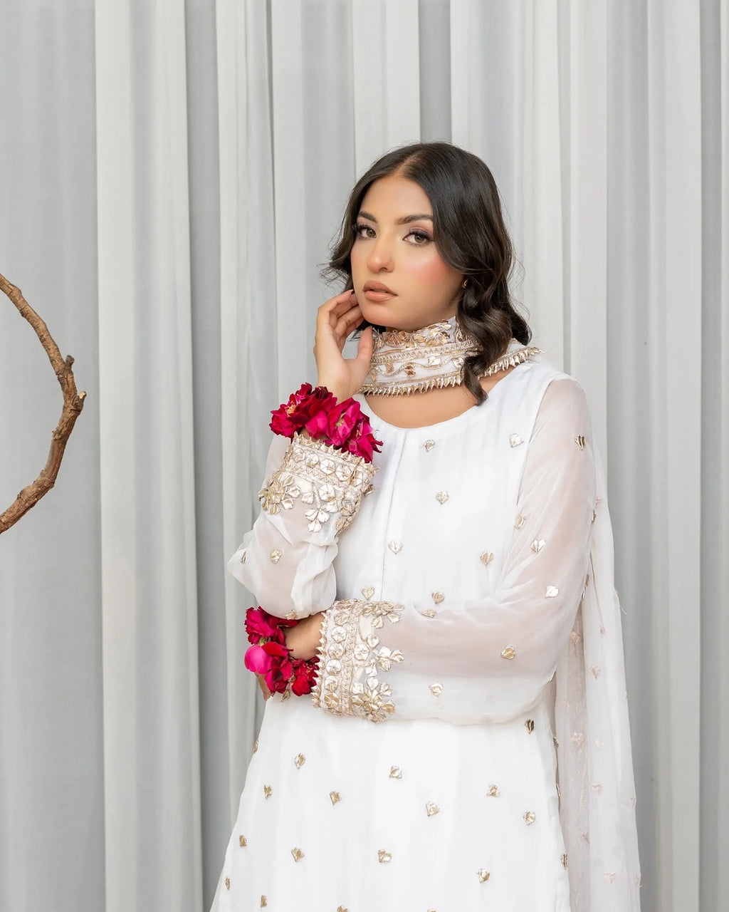 Woman wearing a white and gold embroidered outfit with pink flowers against a gray curtain background
