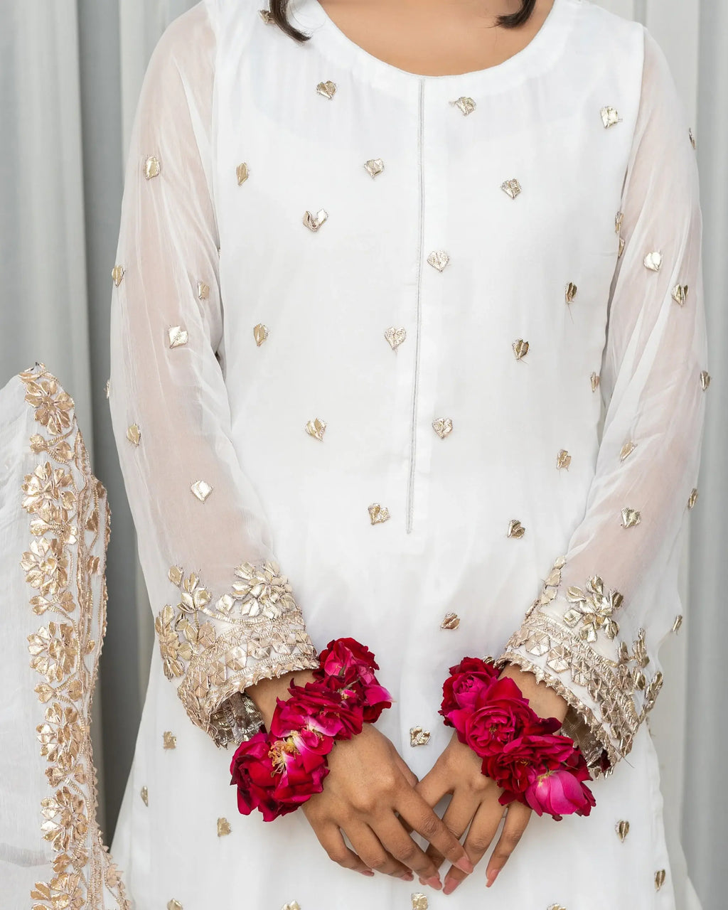 Person wearing a white embroidered outfit with gold details, holding pink flowers.
