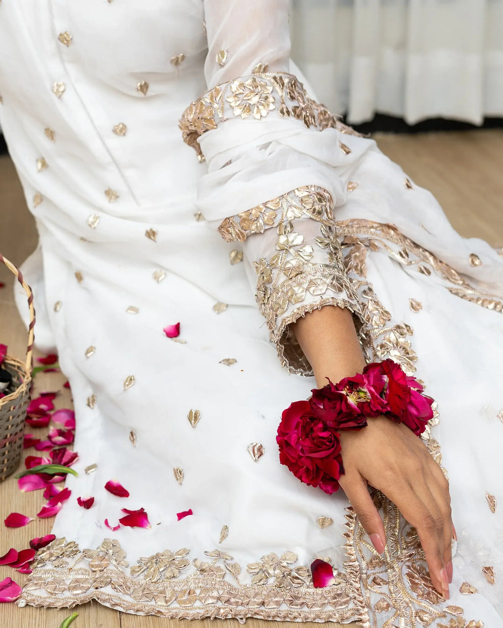White saree with gold embroidery and a hand wearing a red flower bracelet on a wooden surface.