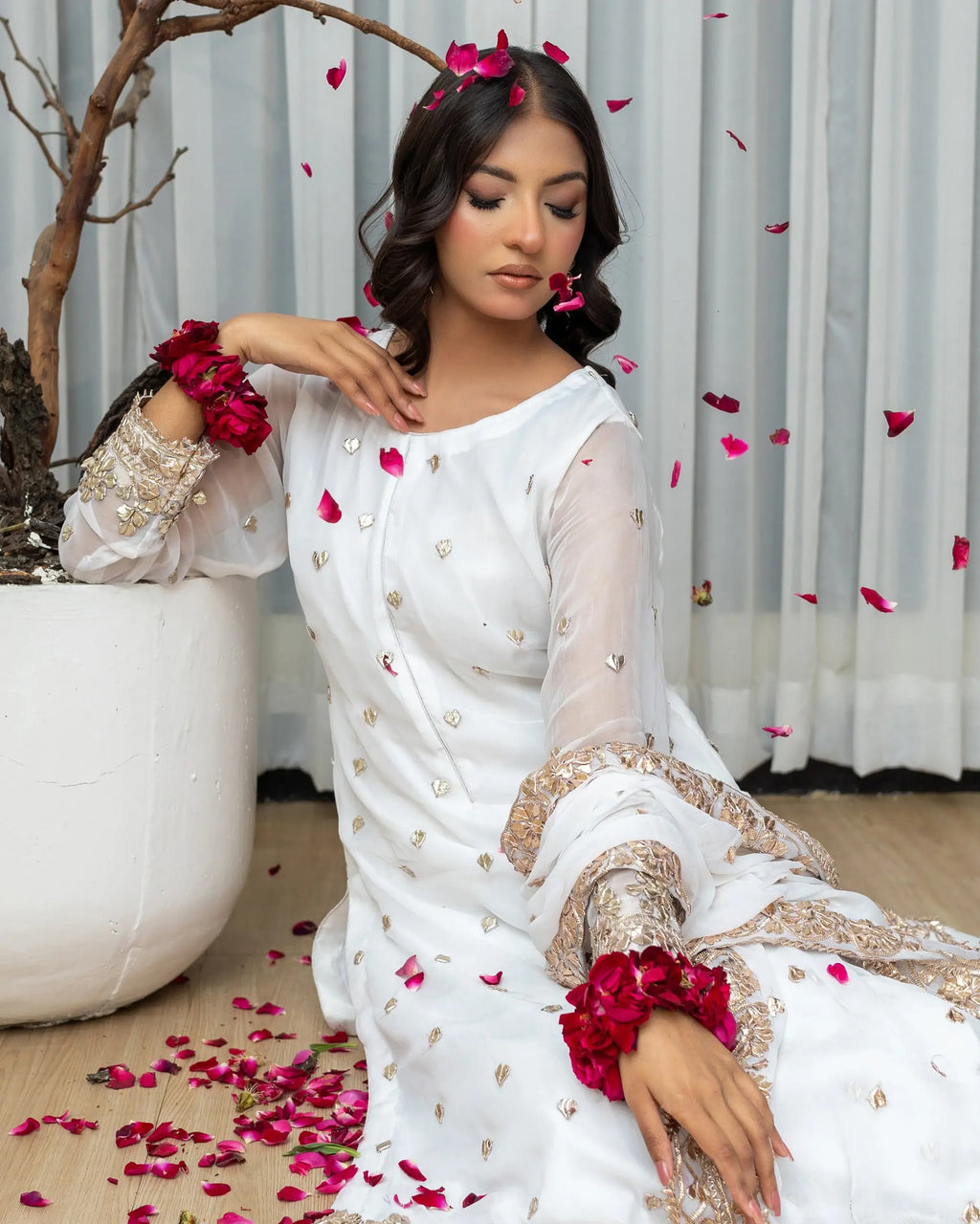 Woman in a white traditional outfit with gold details, surrounded by red flowers and petals.