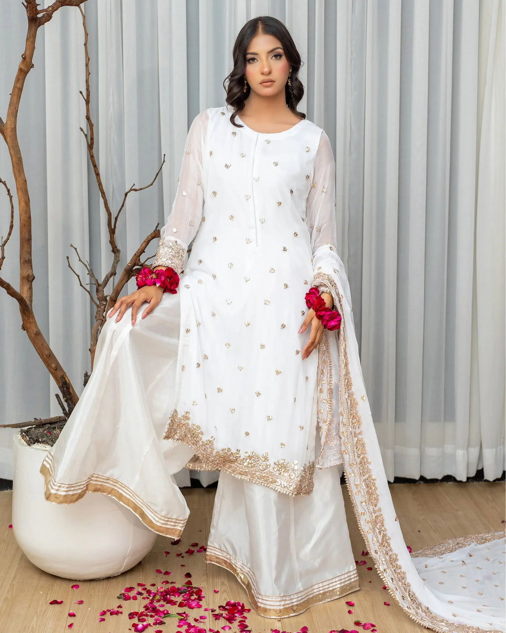 Woman in a white traditional outfit with gold details, standing indoors with a neutral background.