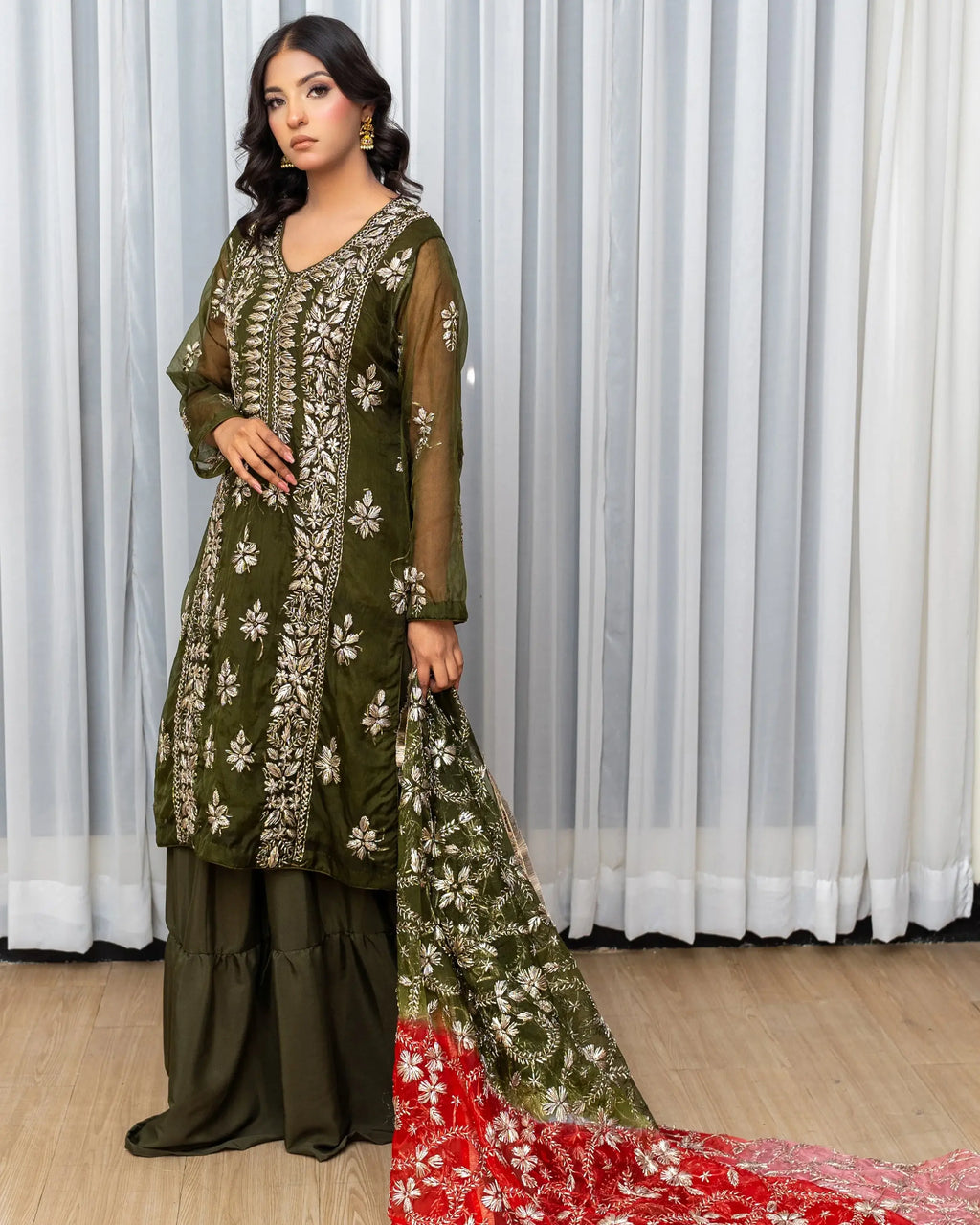 Woman in a green embroidered outfit with a red dupatta against a white curtain background