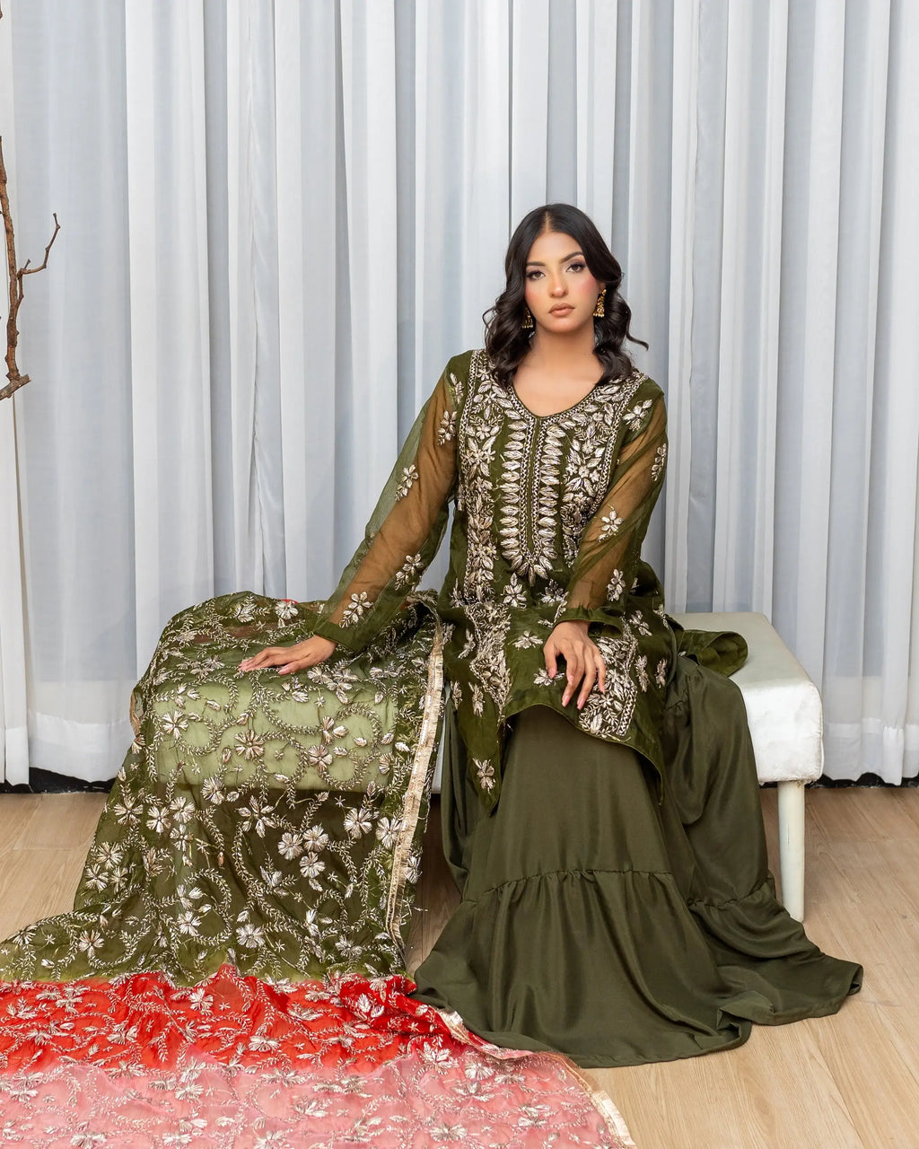 Woman in a green embroidered traditional outfit sitting on a white bench with a white curtain background.