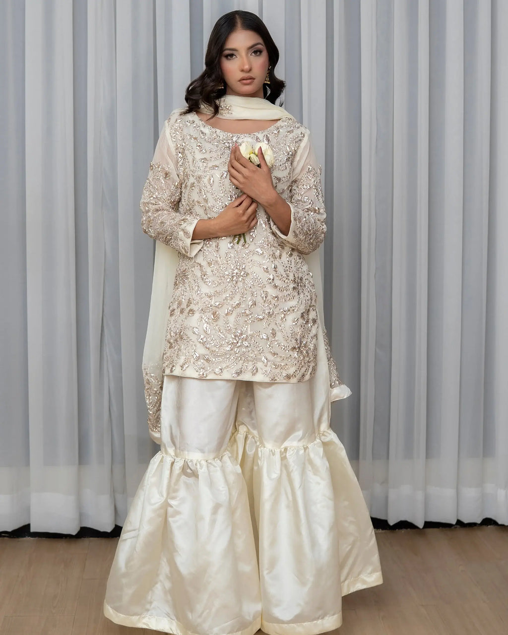 Woman wearing a traditional outfit with a lace top and white pants against a striped curtain background