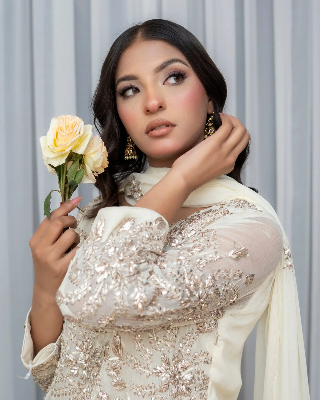 Woman in an embellished outfit holding flowers against a neutral background
