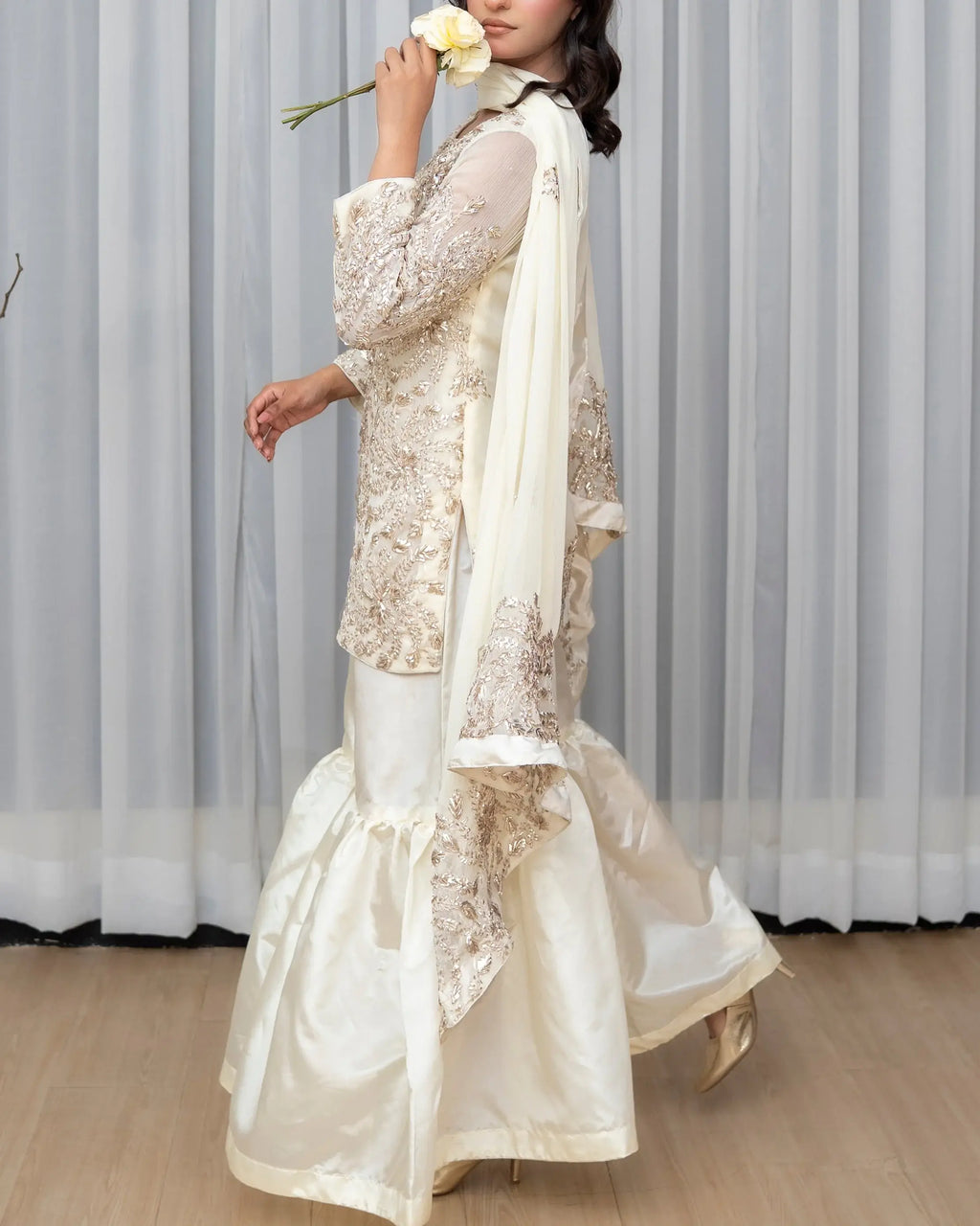 Woman in a white traditional outfit holding a flower against a gray curtain background