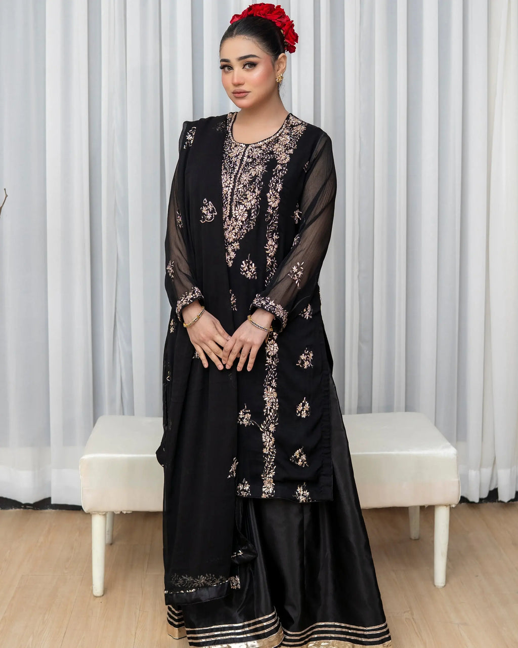 Woman in a black embroidered traditional outfit standing against a white curtain background.