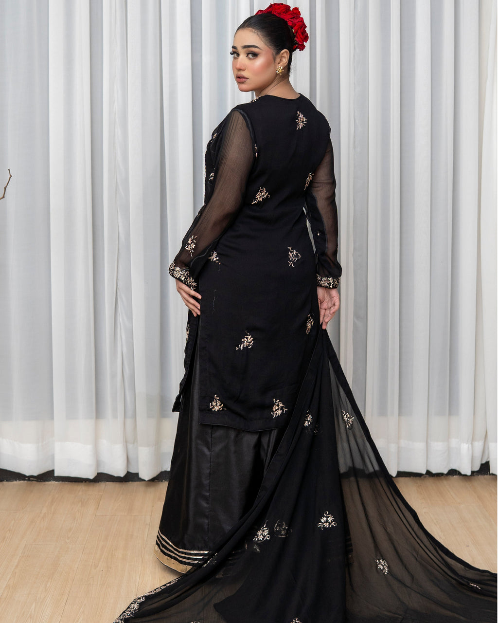 Woman wearing a black saree with floral embroidery in front of white curtains