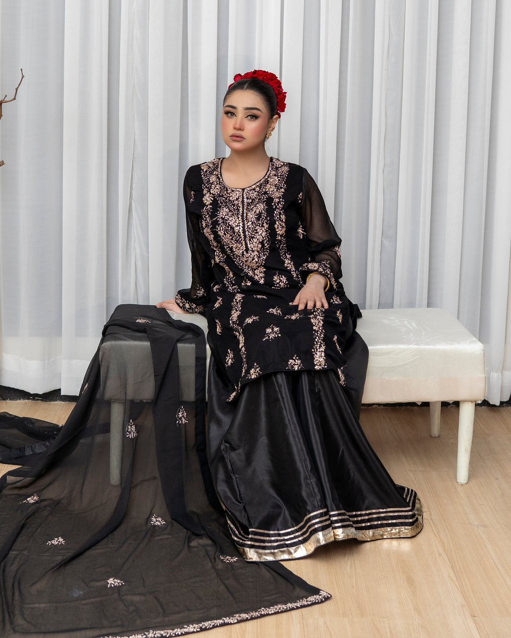 Woman in a black embroidered traditional outfit sitting on a chair with a white curtain background