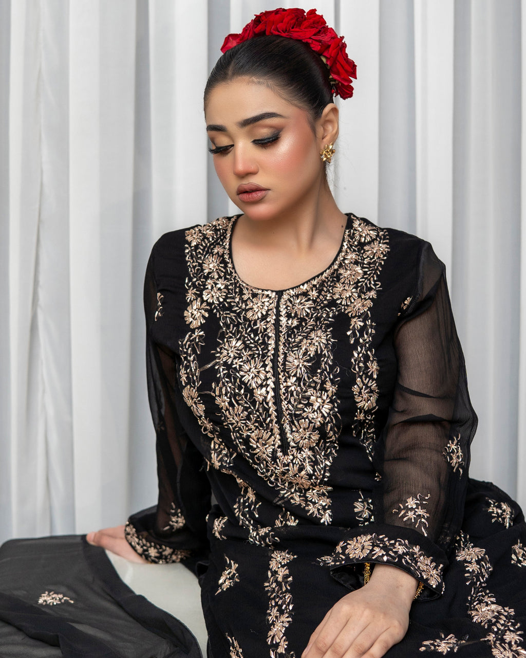Woman wearing a black and gold embroidered outfit with a red flower in her hair against a white curtain background.