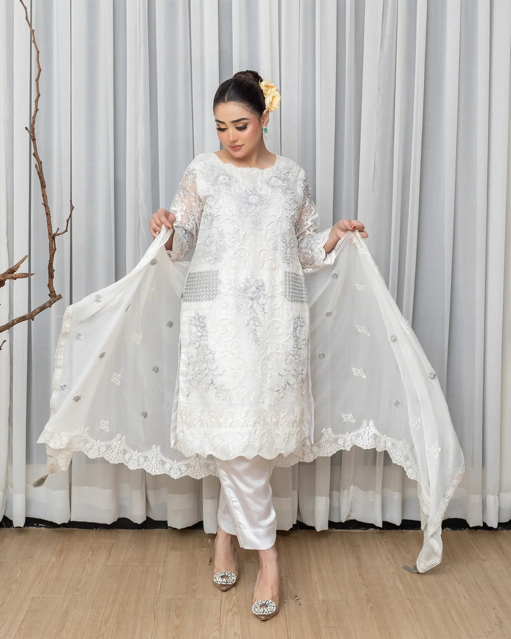 Woman in a white embroidered outfit holding a dupatta against a gray curtain background