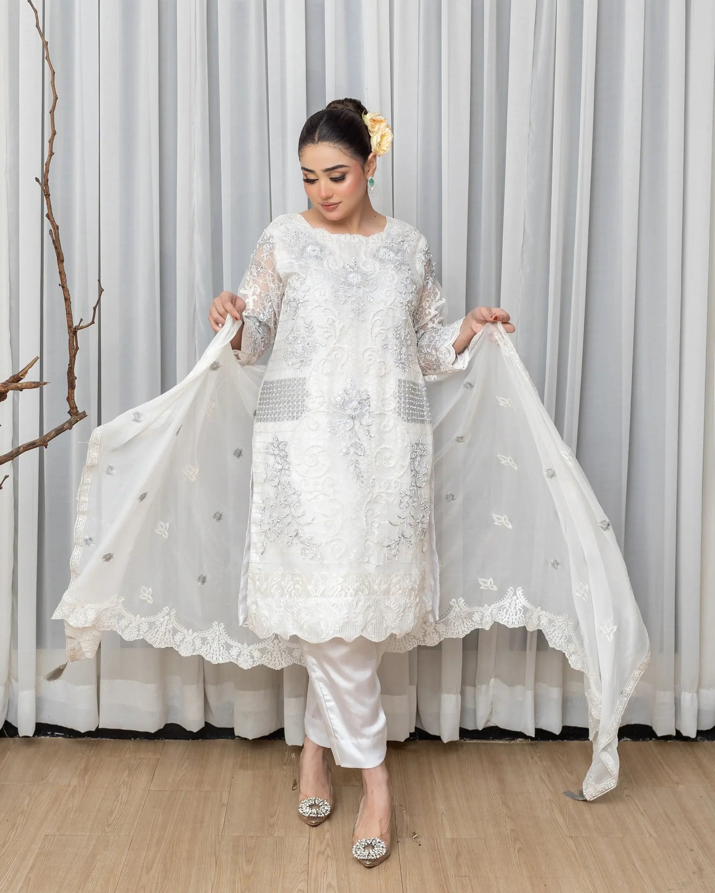 Woman in a white embroidered outfit holding a dupatta against a gray curtain background