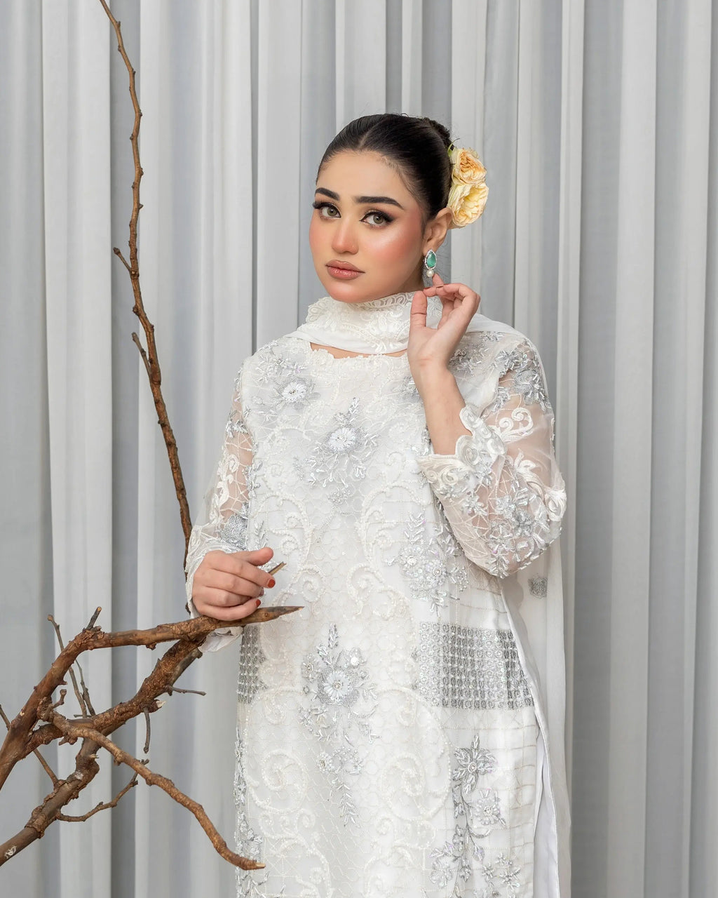 Woman in a white lace dress standing against a gray curtain with decorative branches.