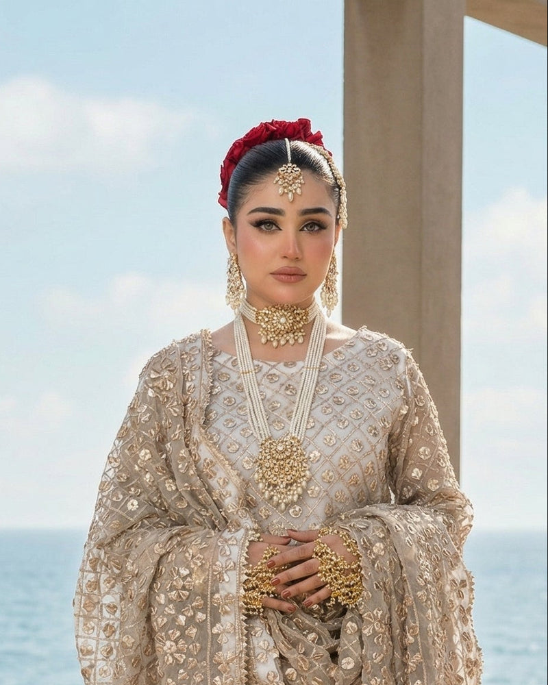 Woman in traditional embroidered outfit with jewelry, standing on a deck with ocean view.