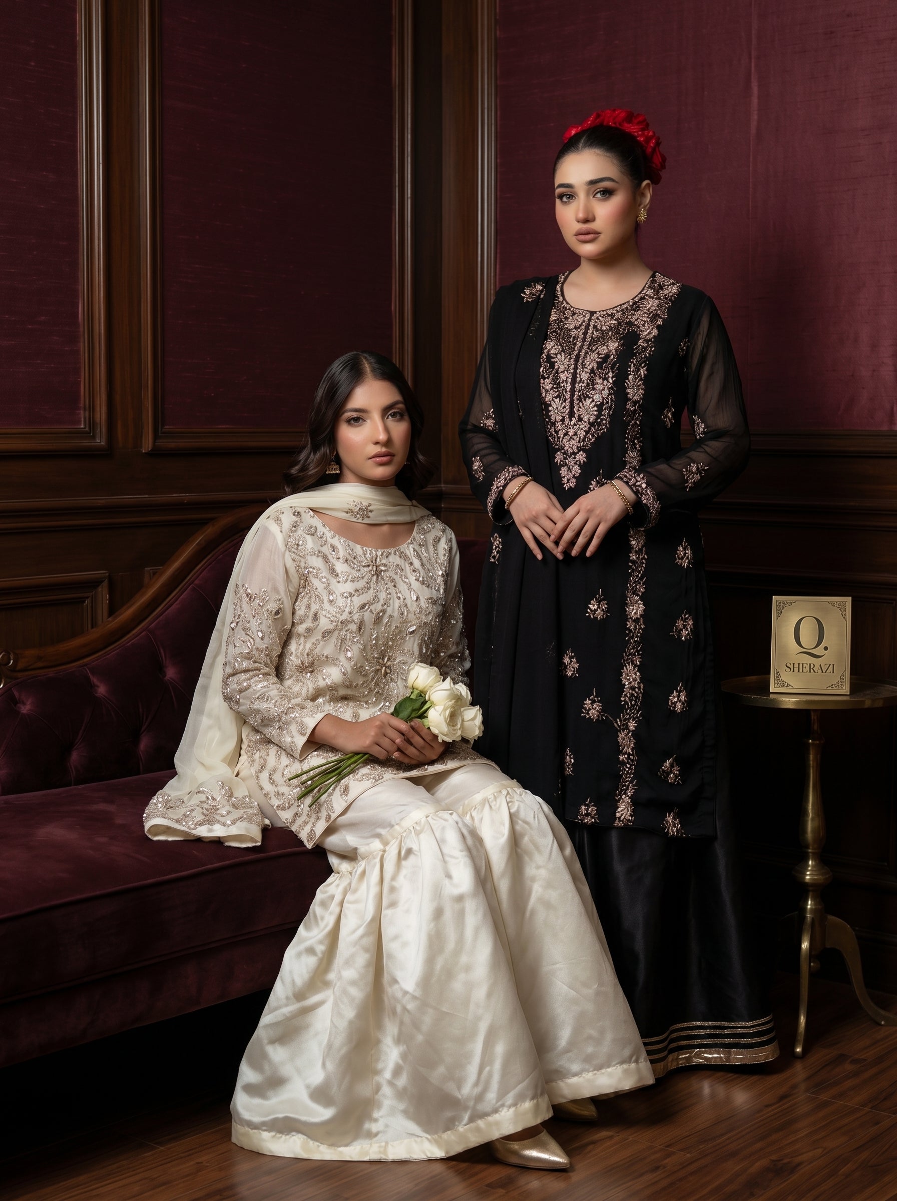 Two women in traditional attire sitting on a couch against a dark red wall.